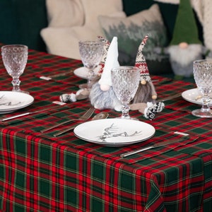 May include: A festive table setting with a red and green plaid tablecloth. Each place setting includes a white plate with a black and white design, silverware, and a crystal wine glass. Decorative gnomes add a whimsical touch.