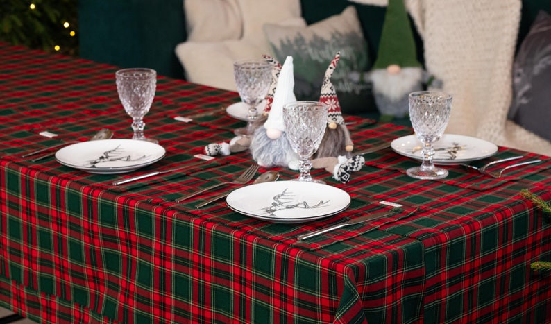 May include: A festive table setting with a red and green plaid tablecloth. Each place setting includes a white plate with a black and white design, silverware, and a crystal wine glass. Decorative gnomes add a whimsical touch.