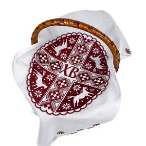 May include: A white cloth with a red embroidered design featuring deer, snowflakes, and geometric patterns, placed inside a woven basket with a wooden handle. The cloth has the letters "XB" embroidered on it.