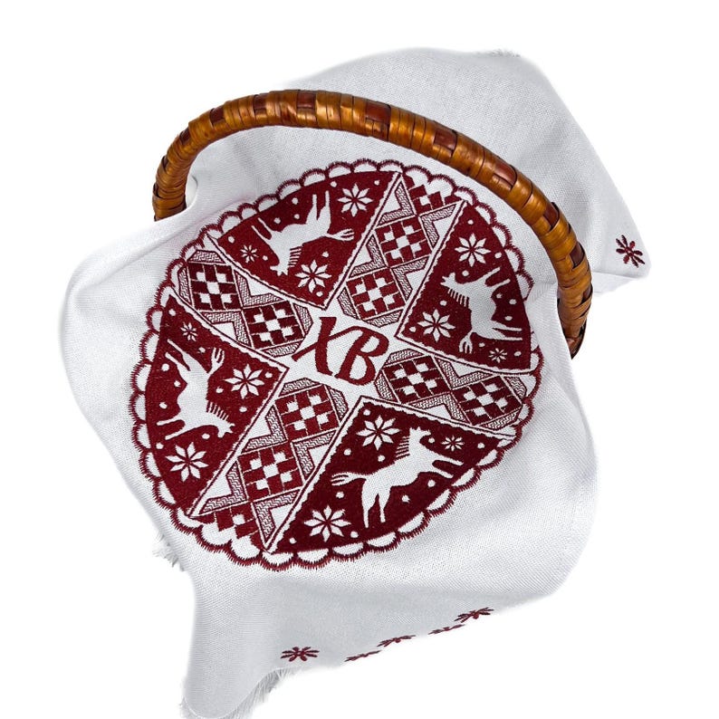 May include: A white cloth with a red embroidered design featuring deer, snowflakes, and geometric patterns, placed inside a woven basket with a wooden handle. The cloth has the letters "XB" embroidered on it.