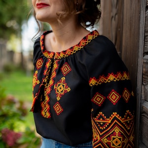 May include: A black embroidered blouse with a red, yellow, and orange geometric pattern. The blouse has a loose fit and a round neckline.