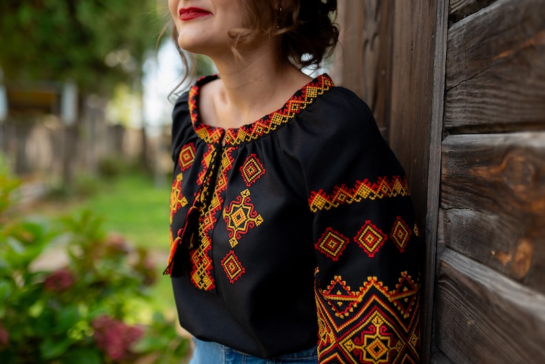 May include: A black embroidered blouse with a red, yellow, and orange geometric pattern. The blouse has a loose fit and a round neckline.
