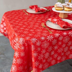 May include: A red tablecloth with a white snowflake pattern. The table is set with plates, napkins, and gold-colored cutlery. A cake stand with pastries is visible on the table.