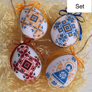 May include: A set of four decorative Easter egg ornaments. Each egg features intricate cross-stitch embroidery in various colors, including blue, orange, and red, on a white background. The eggs are adorned with colorful ribbons and hanging strings.