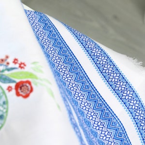 May include: White fabric with a blue geometric border and floral embroidery. The border features a repeating pattern of blue diamonds and zigzags. Red and green floral embroidery is visible on the left side of the fabric.