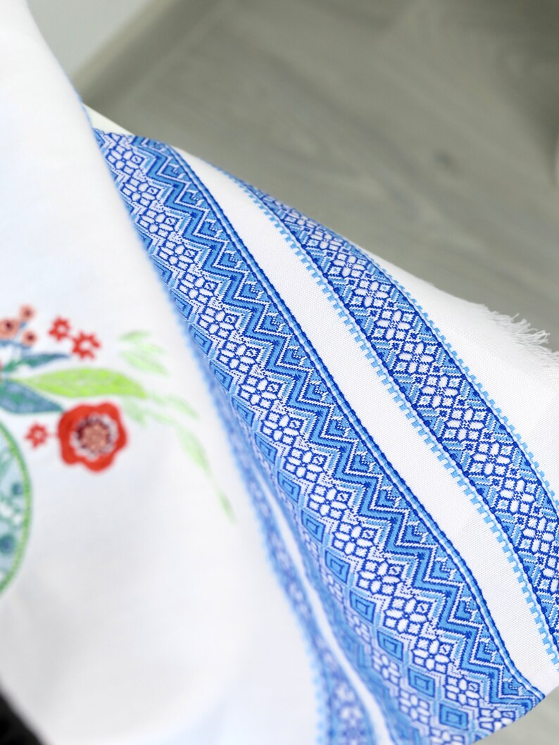 May include: White fabric with a blue geometric border and floral embroidery. The border features a repeating pattern of blue diamonds and zigzags. Red and green floral embroidery is visible on the left side of the fabric.