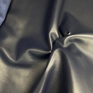 May include: A close-up of a dark blue leather fabric with a smooth, textured surface. The fabric is folded and draped, showcasing its soft and supple texture.