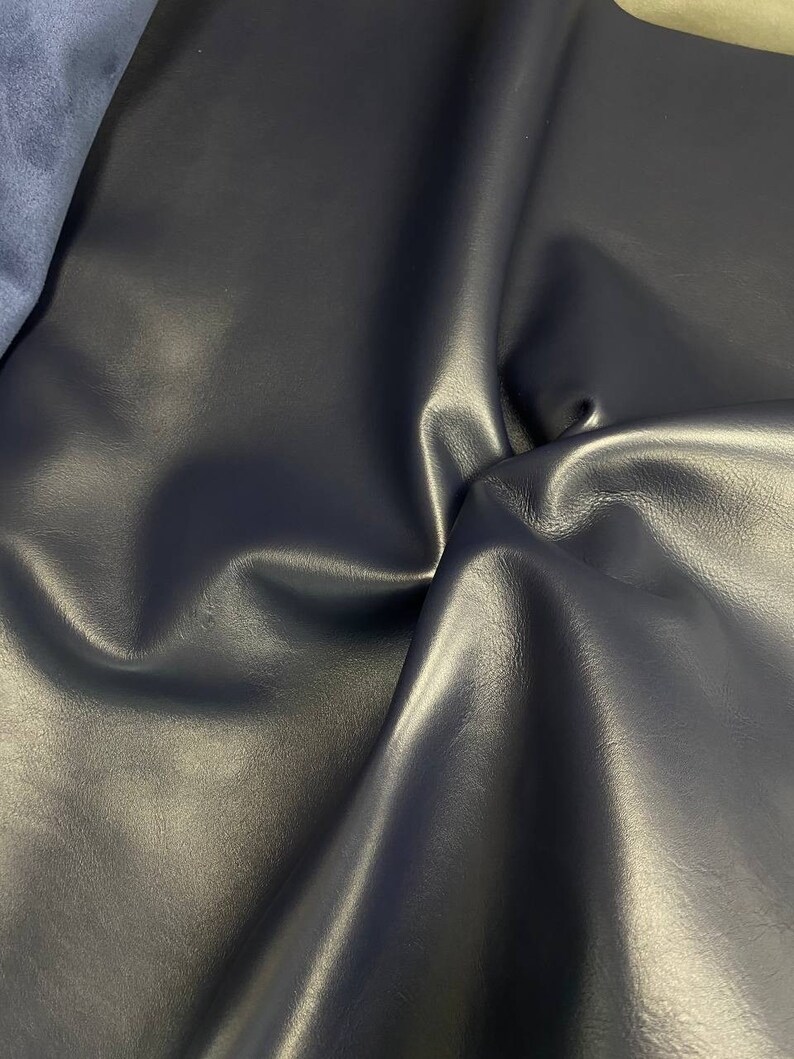 May include: A close-up of a dark blue leather fabric with a smooth, textured surface. The fabric is folded and draped, showcasing its soft and supple texture.