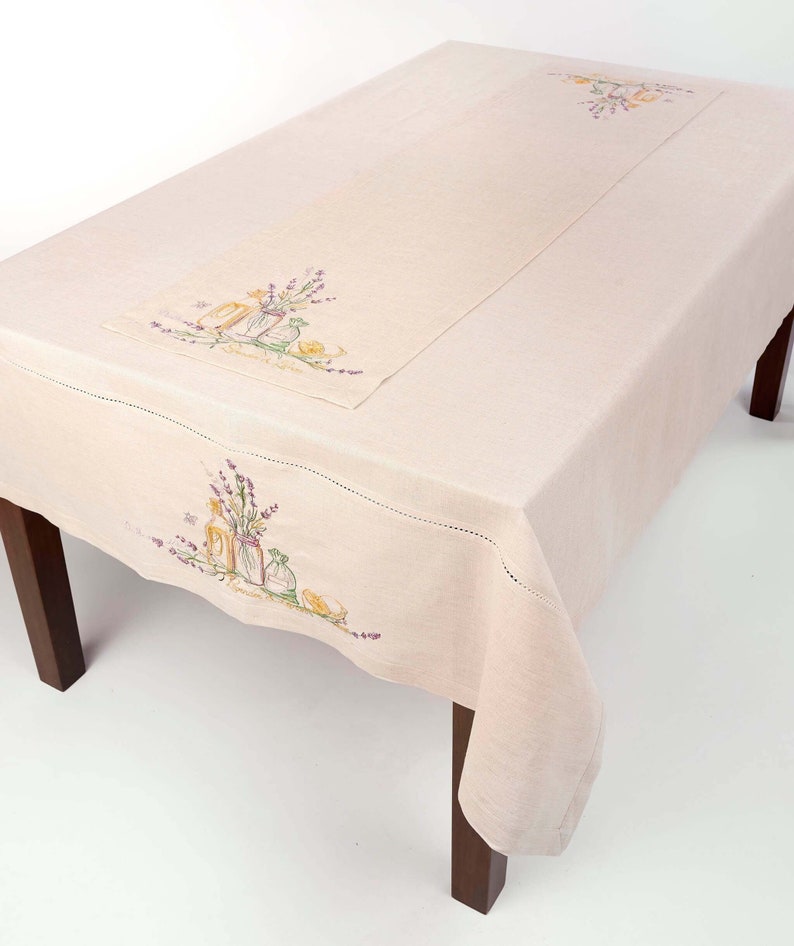 May include: A light beige linen tablecloth with a matching runner. Both feature embroidered floral designs in purple, yellow, and green, depicting vases and sprigs of lavender. The tablecloth is draped over a dark wooden table.