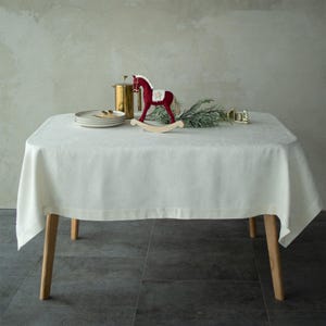 Op de afbeelding: Een witte tafelkleed bedekt een tafel met houten poten, versierd met een rood schommelpaard, borden en een goudkleurige container. De tafelsetting bevat een takje groen, waardoor een feestelijk vakantiethema ontstaat.