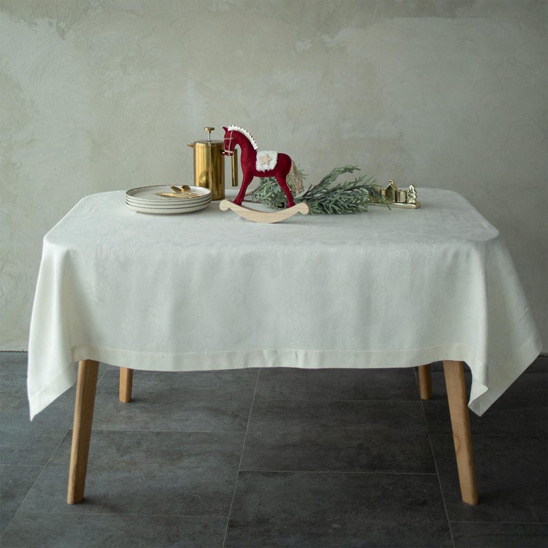 Op de afbeelding: Een witte tafelkleed bedekt een tafel met houten poten, versierd met een rood schommelpaard, borden en een goudkleurige container. De tafelsetting bevat een takje groen, waardoor een feestelijk vakantiethema ontstaat.