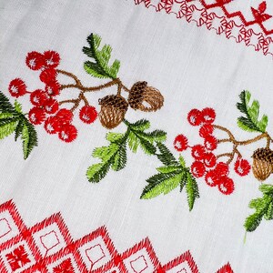 K&ouml;nnte beinhalten: Wei&szlig;er Stoff mit Stickereien. Mit roten Beeren, braunen Eicheln und gr&uuml;nen Bl&auml;ttern. Rote geometrische und florale Muster s&auml;umen das Design. Die Stickerei ist detailliert und lebendig und erzeugt einen strukturierten Look.