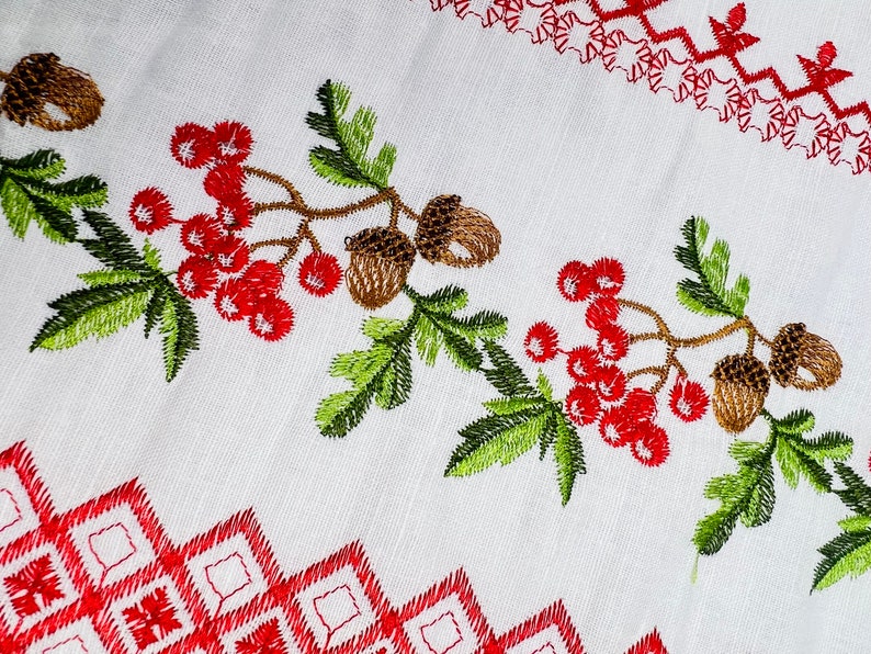 K&ouml;nnte beinhalten: Wei&szlig;er Stoff mit Stickereien. Mit roten Beeren, braunen Eicheln und gr&uuml;nen Bl&auml;ttern. Rote geometrische und florale Muster s&auml;umen das Design. Die Stickerei ist detailliert und lebendig und erzeugt einen strukturierten Look.