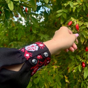K&ouml;nnte beinhalten: Schwarze Bluse mit langen &Auml;rmeln und roten und wei&szlig;en Stickereien an den Manschetten. Die Manschetten haben zwei schwarze Kn&ouml;pfe. Die Bluse wird vor einem Hintergrund aus gr&uuml;nen Bl&auml;ttern und roten Beeren gezeigt.