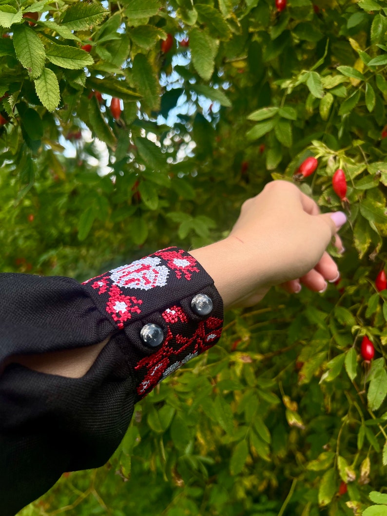 K&ouml;nnte beinhalten: Schwarze Bluse mit langen &Auml;rmeln und roten und wei&szlig;en Stickereien an den Manschetten. Die Manschetten haben zwei schwarze Kn&ouml;pfe. Die Bluse wird vor einem Hintergrund aus gr&uuml;nen Bl&auml;ttern und roten Beeren gezeigt.