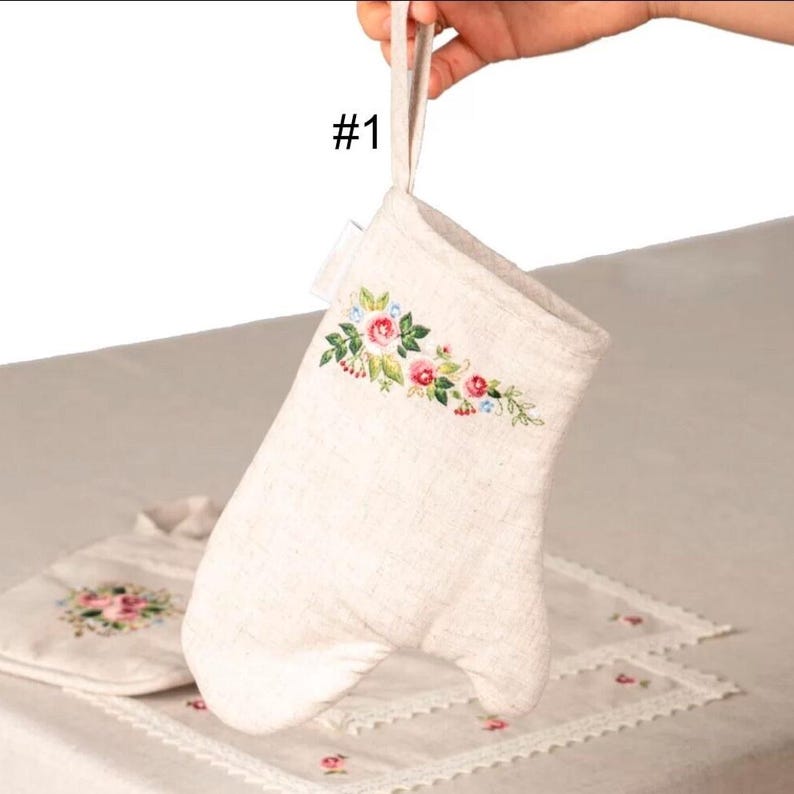 May include: Off-white oven mitt with a floral embroidery design in pink, red, and blue. The mitt has a loop for hanging and is part of a kitchen set. The text "#1" is visible.