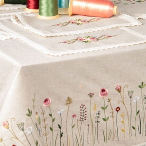 May include: A beige linen tablecloth with embroidered wildflowers and a lace trim. Several spools of thread in various colors, including green, red, and pink, are arranged on the table, along with matching embroidered placemats.