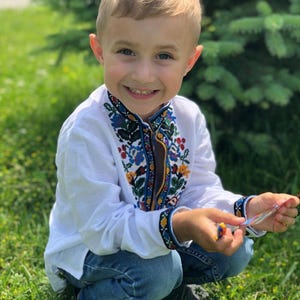 May include: A young boy wearing a white embroidered shirt with blue, yellow, red, and green floral patterns. He is wearing blue jeans and is smiling at the camera.