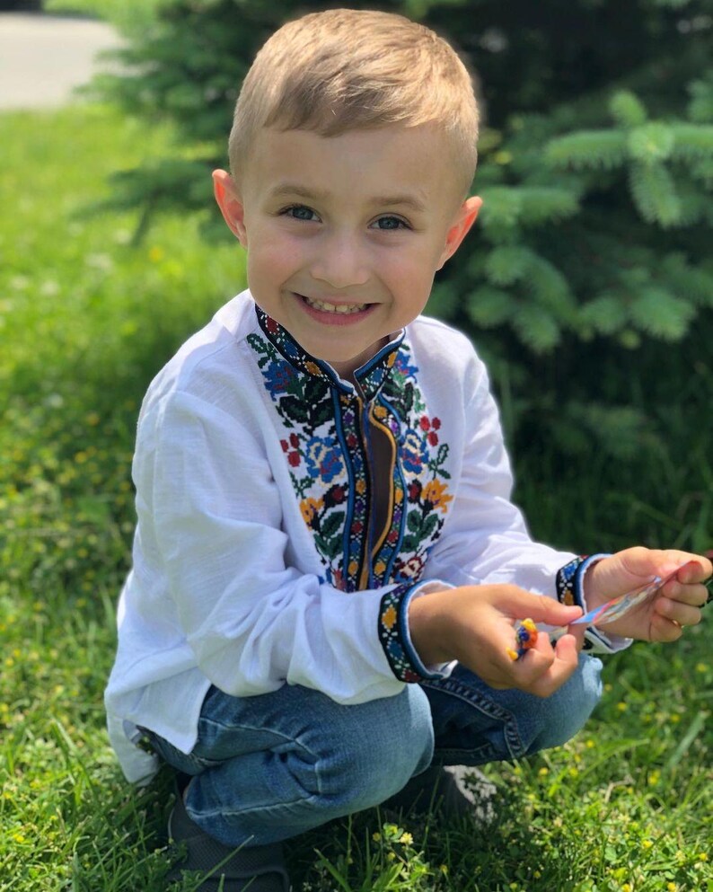 May include: A young boy wearing a white embroidered shirt with blue, yellow, red, and green floral patterns. He is wearing blue jeans and is smiling at the camera.