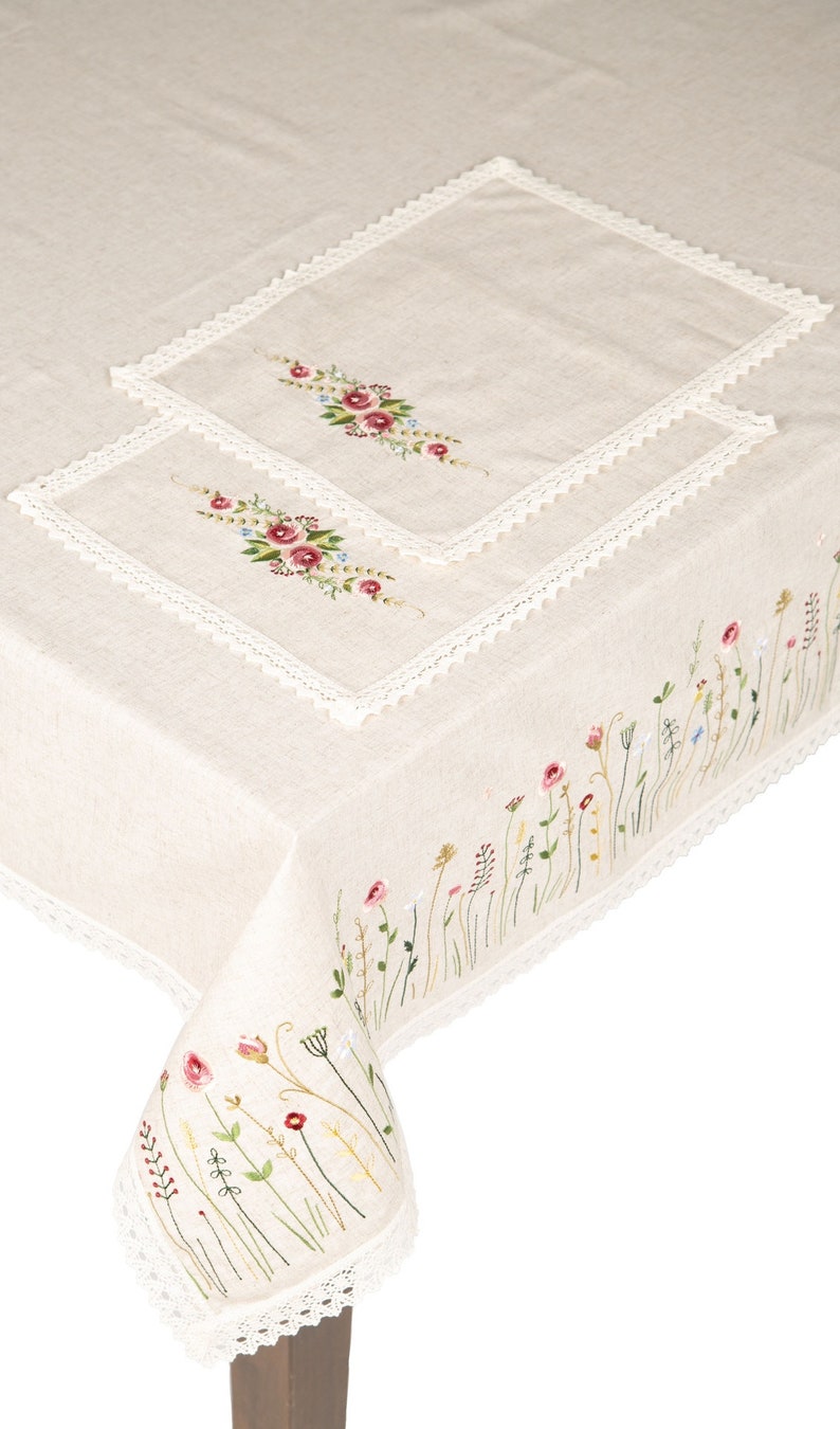 May include: A white linen tablecloth with delicate floral embroidery. The tablecloth features a lace trim and a smaller embroidered square in the center. The embroidery is in shades of pink, green, and blue.