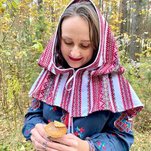 May include: A woman wearing a traditional embroidered hood and dress. The hood is white with red geometric patterns. The dress is navy blue with colorful floral embroidery on the sleeves and bodice. The woman is holding a mushroom.
