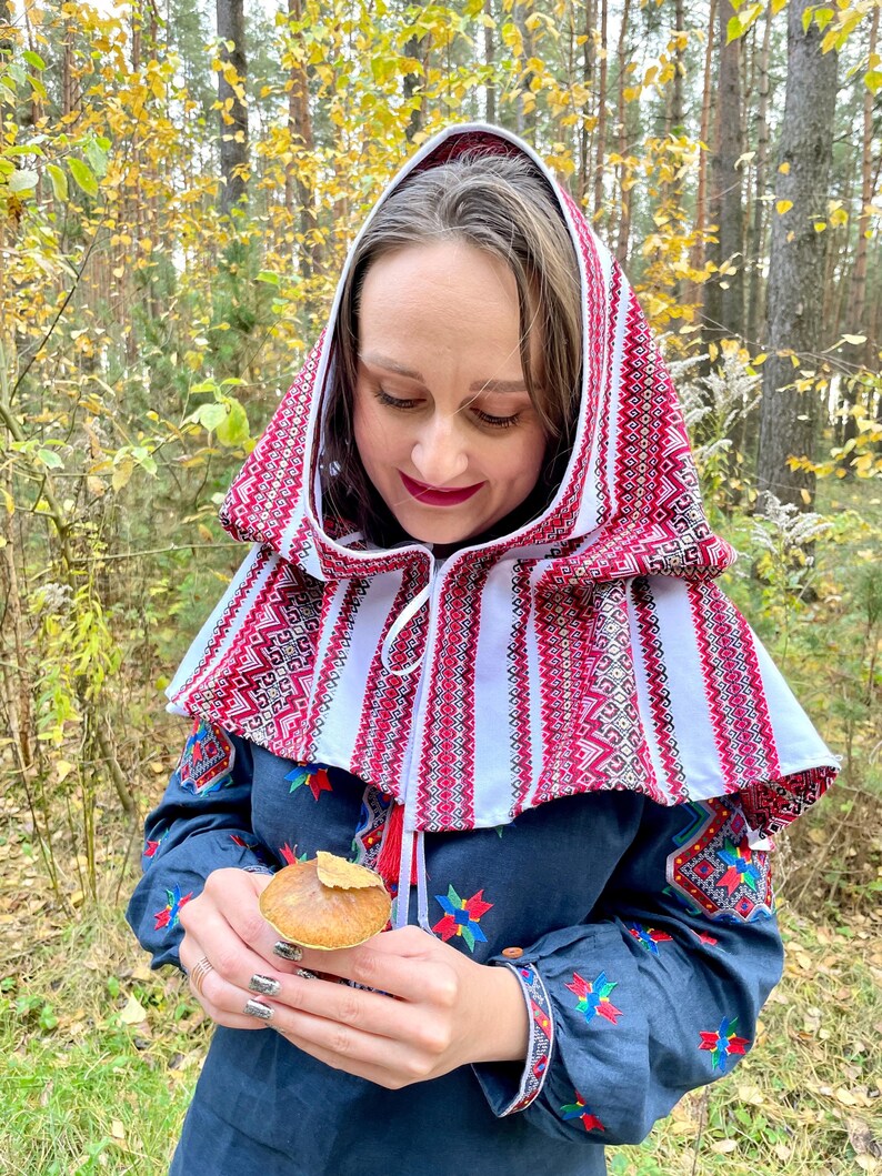 May include: A woman wearing a traditional embroidered hood and dress. The hood is white with red geometric patterns. The dress is navy blue with colorful floral embroidery on the sleeves and bodice. The woman is holding a mushroom.