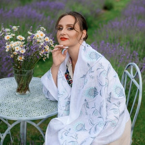 Puede incluir: Una mujer con una bufanda blanca con un estampado floral, sentada en una mesa peque&ntilde;a con un jarr&oacute;n de margaritas. El fondo presenta un campo de lavanda y una bicicleta blanca decorativa.