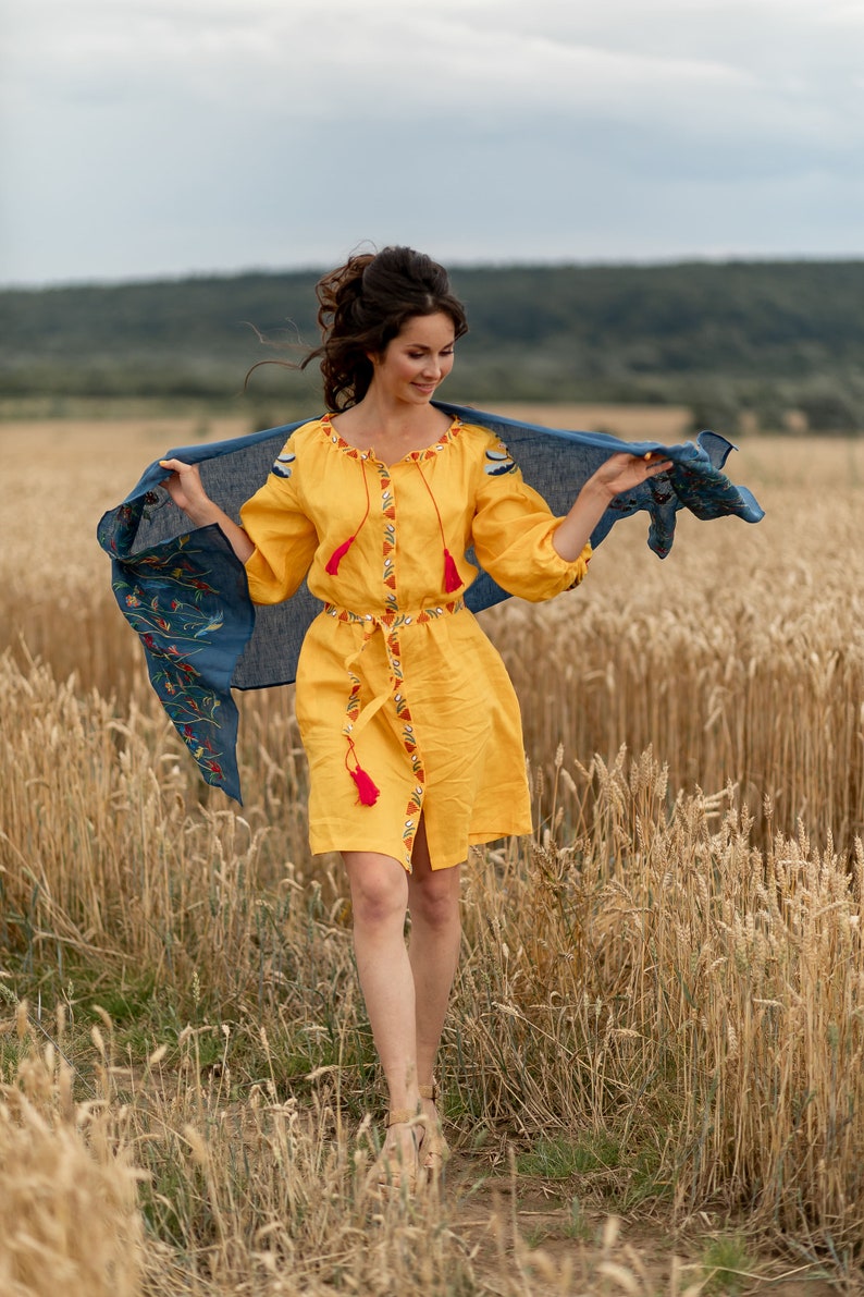 May include: A woman in a yellow embroidered dress with a blue scarf walks through a field of wheat.