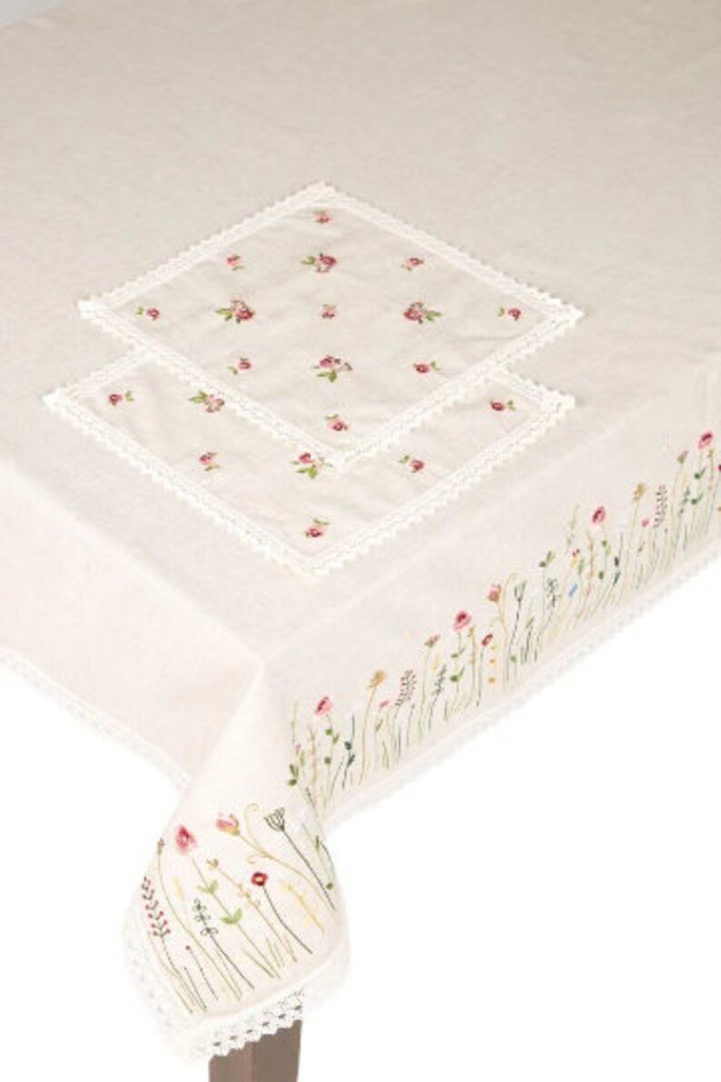 May include: Cream-colored tablecloth with embroidered floral details. Features a border of wildflowers and two matching square placemats with small pink flower embroidery and lace trim. A delicate and elegant design.