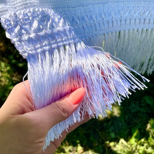 Puede incluir: Primer plano de un textil blanco bordado con detalles florales rojos. La tela presenta un patr&oacute;n de punto de cruz detallado y un borde de flecos. El textil se sostiene en una mano, mostrando la delicada flecos.