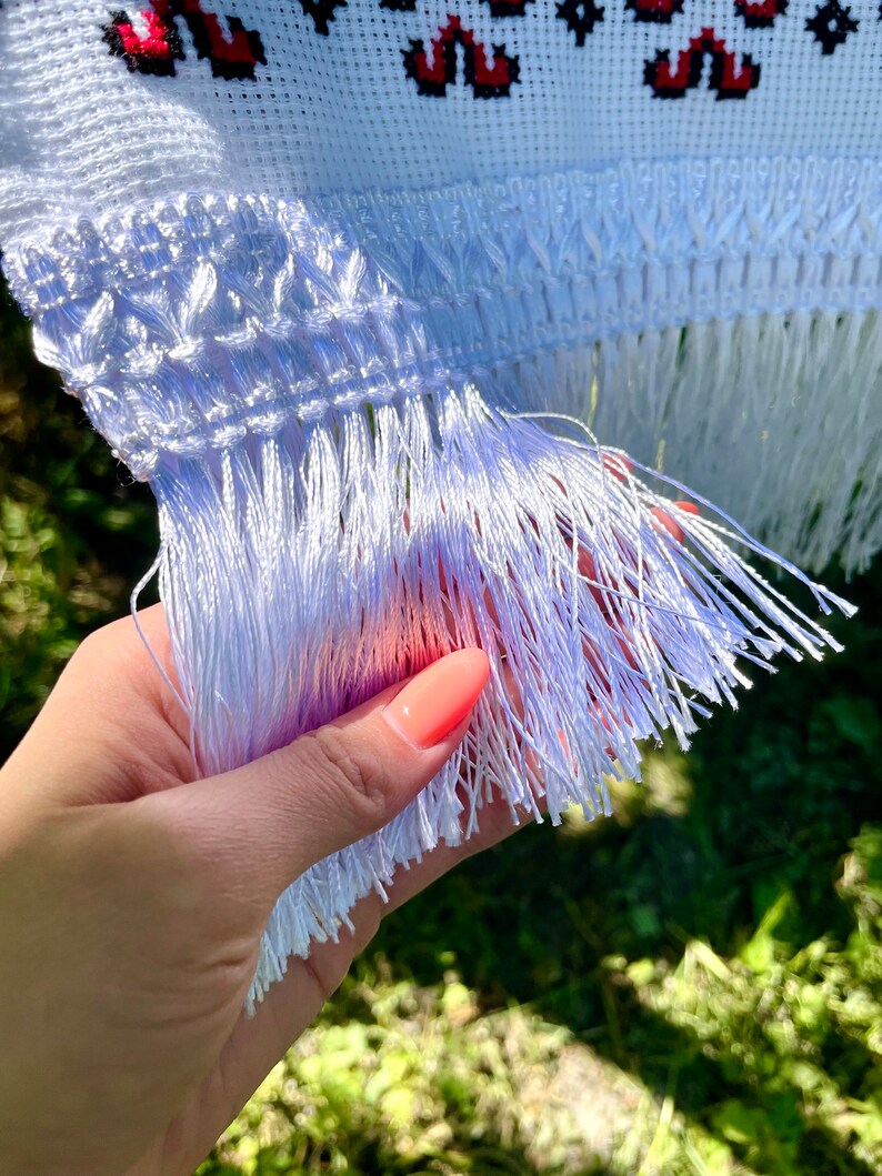 Puede incluir: Primer plano de un textil blanco bordado con detalles florales rojos. La tela presenta un patr&oacute;n de punto de cruz detallado y un borde de flecos. El textil se sostiene en una mano, mostrando la delicada flecos.