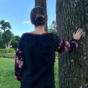 K&ouml;nnte beinhalten: Eine schwarze bestickte Bluse mit roten und wei&szlig;en Blumenmustern an den &Auml;rmeln. Die Bluse hat einen runden Ausschnitt und wird von einer Person getragen, die vor einem Baum steht.
