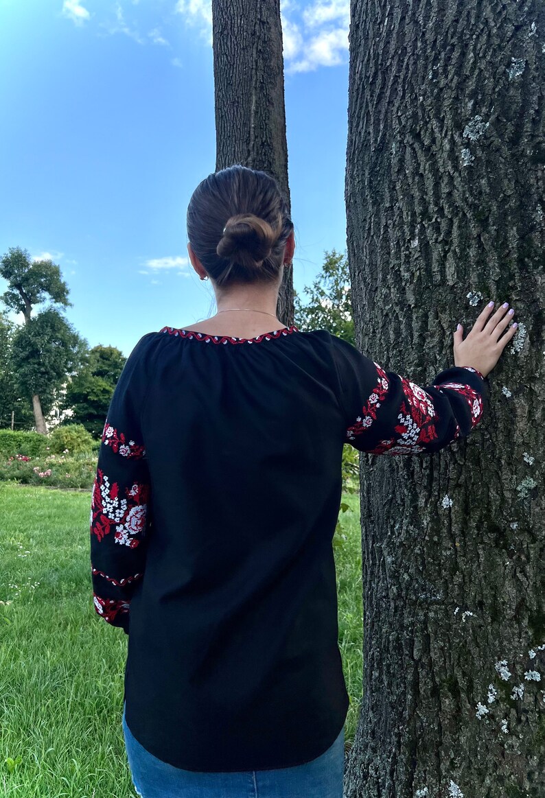 K&ouml;nnte beinhalten: Eine schwarze bestickte Bluse mit roten und wei&szlig;en Blumenmustern an den &Auml;rmeln. Die Bluse hat einen runden Ausschnitt und wird von einer Person getragen, die vor einem Baum steht.