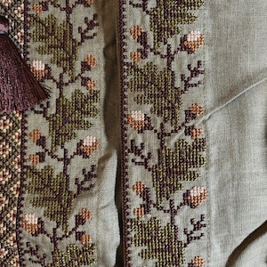 以下が含まれることがあります： オリーブグリーンの生地に、詳細なクロスステッチ刺繍が施されたクローズアップ。刺繍は、茶色、緑、クリーム色の花柄のデザインです。左側には、装飾的な要素を加える濃い茶色のタッセルが見えます。