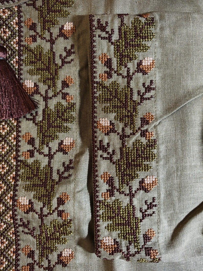 以下が含まれることがあります： オリーブグリーンの生地に、詳細なクロスステッチ刺繍が施されたクローズアップ。刺繍は、茶色、緑、クリーム色の花柄のデザインです。左側には、装飾的な要素を加える濃い茶色のタッセルが見えます。