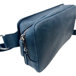 May include: A dark blue leather fanny pack with a silver zipper and a silver buckle. The bag has a strap that can be adjusted to fit around the waist.