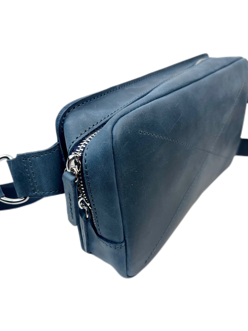 May include: A dark blue leather fanny pack with a silver zipper and a silver buckle. The bag has a strap that can be adjusted to fit around the waist.