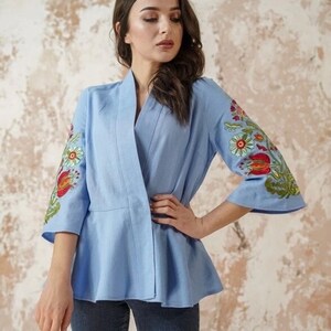 Op de afbeelding: Een lichtblauw linnen blouse met een brede kraag en 3/4 mouwen. De mouwen zijn versierd met kleurrijke geborduurde bloemenmotieven.