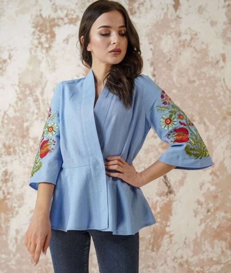 Op de afbeelding: Een lichtblauw linnen blouse met een brede kraag en 3/4 mouwen. De mouwen zijn versierd met kleurrijke geborduurde bloemenmotieven.