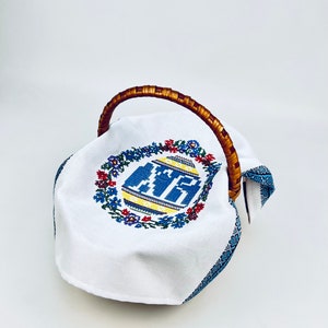 Peut inclure: Un panier de P&acirc;ques en tissu blanc avec un motif d'&oelig;uf brod&eacute; bleu et jaune et une poign&eacute;e en osier brun.