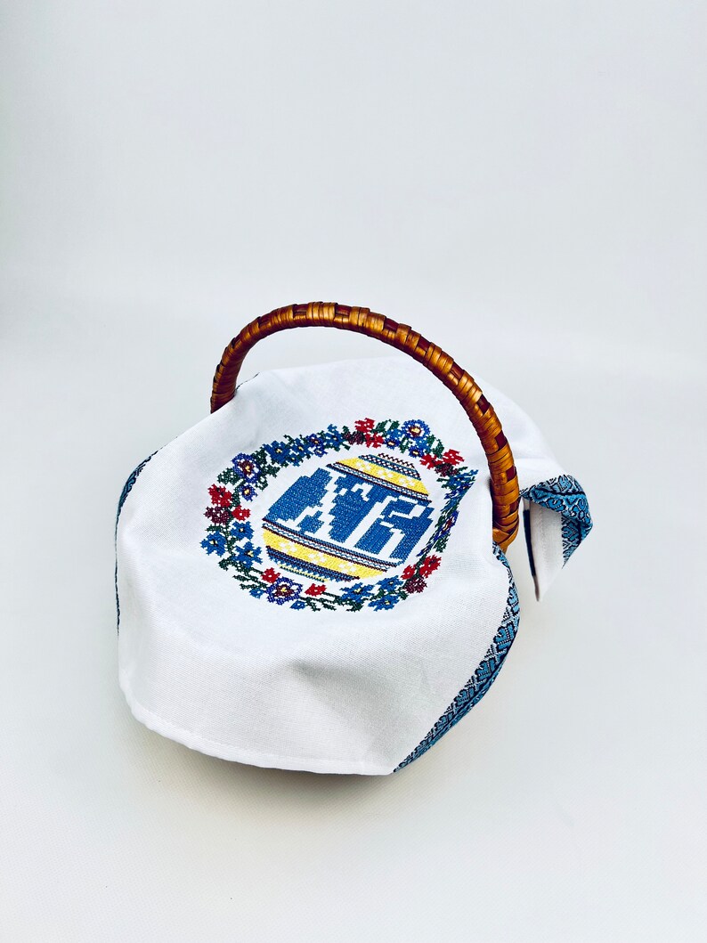 Peut inclure: Un panier de P&acirc;ques en tissu blanc avec un motif d'&oelig;uf brod&eacute; bleu et jaune et une poign&eacute;e en osier brun.
