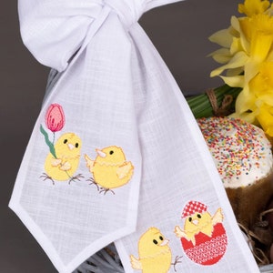 May include: White linen Easter table runner with embroidered yellow chicks and a red egg. One chick is holding a pink tulip. The runner is tied with a white bow.