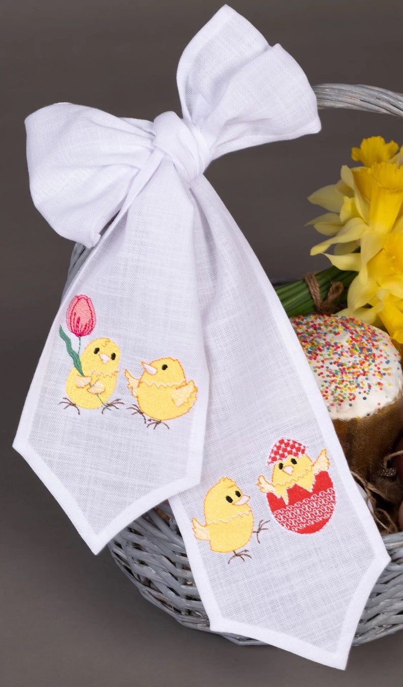 May include: White linen Easter table runner with embroidered yellow chicks and a red egg. One chick is holding a pink tulip. The runner is tied with a white bow.