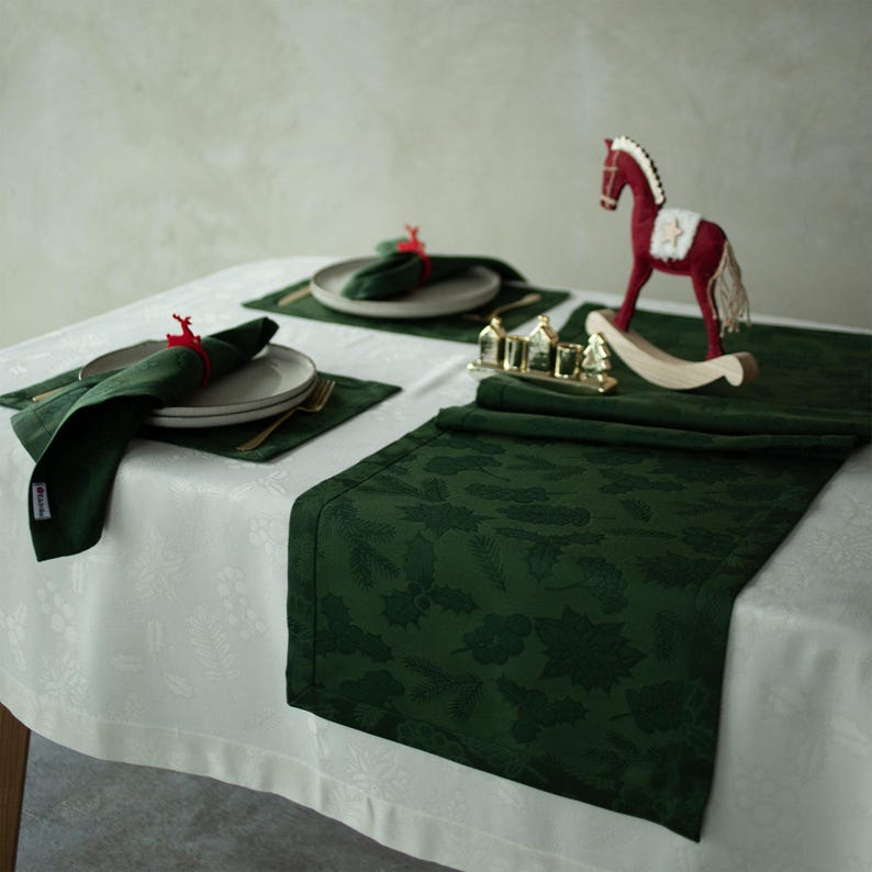 Op de afbeelding: Een feestelijke tafelsetting met een witte tafelkleed met een subtiel bloemenpatroon. Donkergroene placemats, servetten en een tafelloper met een bladerenpatroon zijn geplaatst. Borden, bestek en een rood schommelpaard completeren de decoratie.