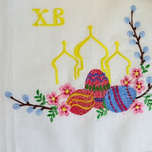 May include: White fabric embroidered with yellow text "XB", three yellow church domes, and colourful Easter eggs. The design includes pink flowers, blue buds, and green leaves, creating a festive Easter theme.
