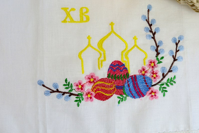 May include: White fabric embroidered with yellow text "XB", three yellow church domes, and colourful Easter eggs. The design includes pink flowers, blue buds, and green leaves, creating a festive Easter theme.