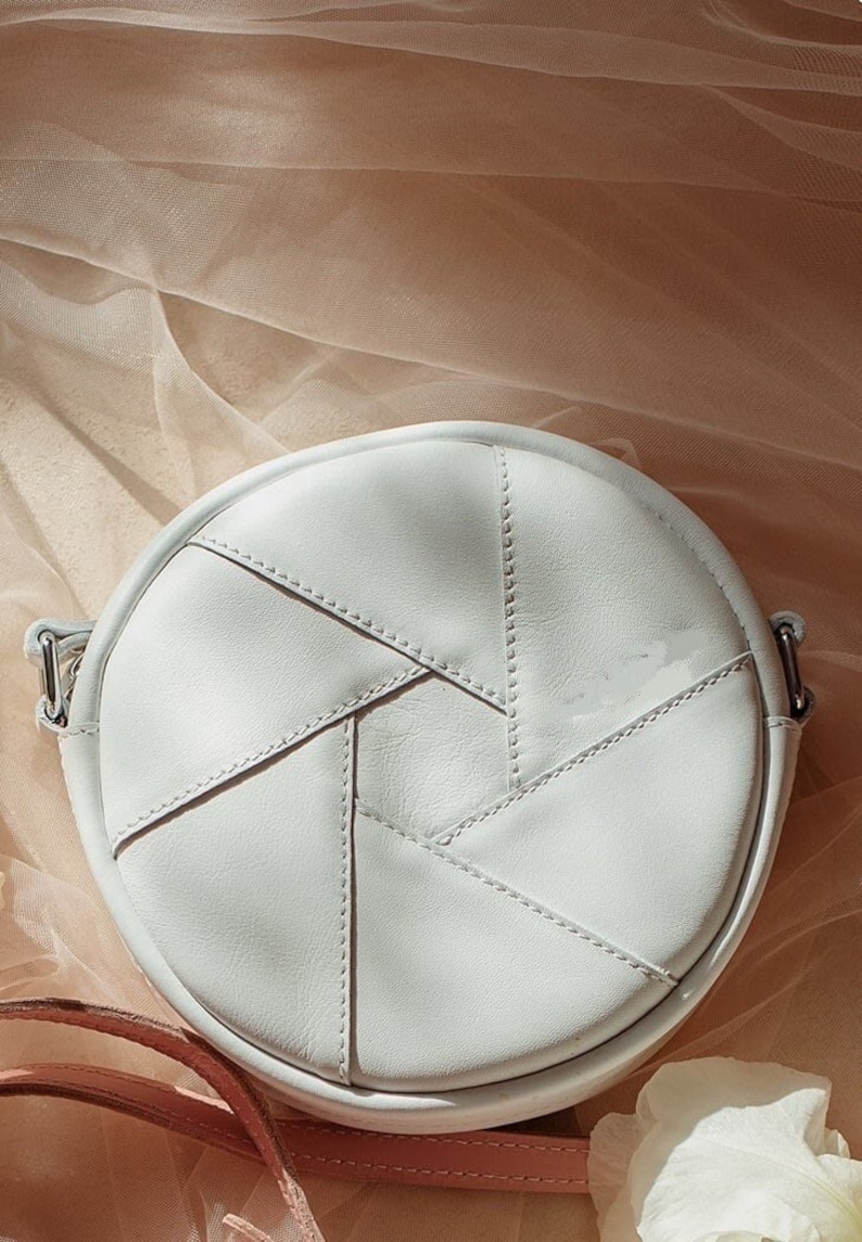 May include: A white round leather crossbody bag with a geometric design. The bag has a single strap and is made of stitched leather panels.