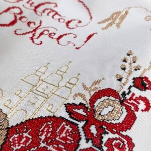Puede incluir: Primer plano de una tela blanca con bordados intrincados. El dise&ntilde;o presenta texto cir&iacute;lico rojo, motivos florales y detalles dorados. El estilo de bordado es detallado y tradicional.