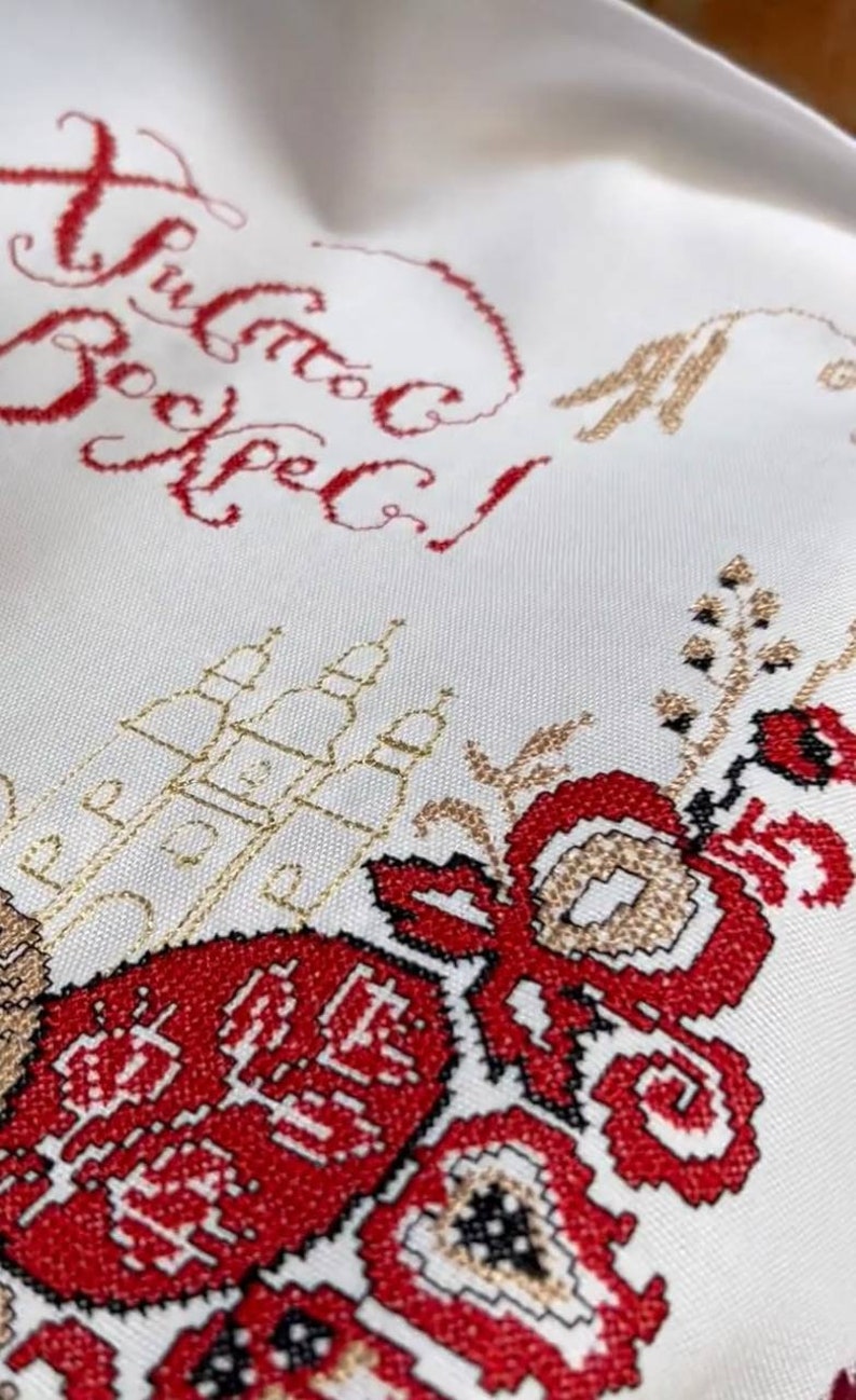 Puede incluir: Primer plano de una tela blanca con bordados intrincados. El dise&ntilde;o presenta texto cir&iacute;lico rojo, motivos florales y detalles dorados. El estilo de bordado es detallado y tradicional.