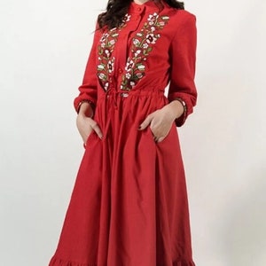May include: A red dress with long sleeves and a ruffled hem. The dress features floral embroidery around the collar and down the front. It has a drawstring waist and pockets.